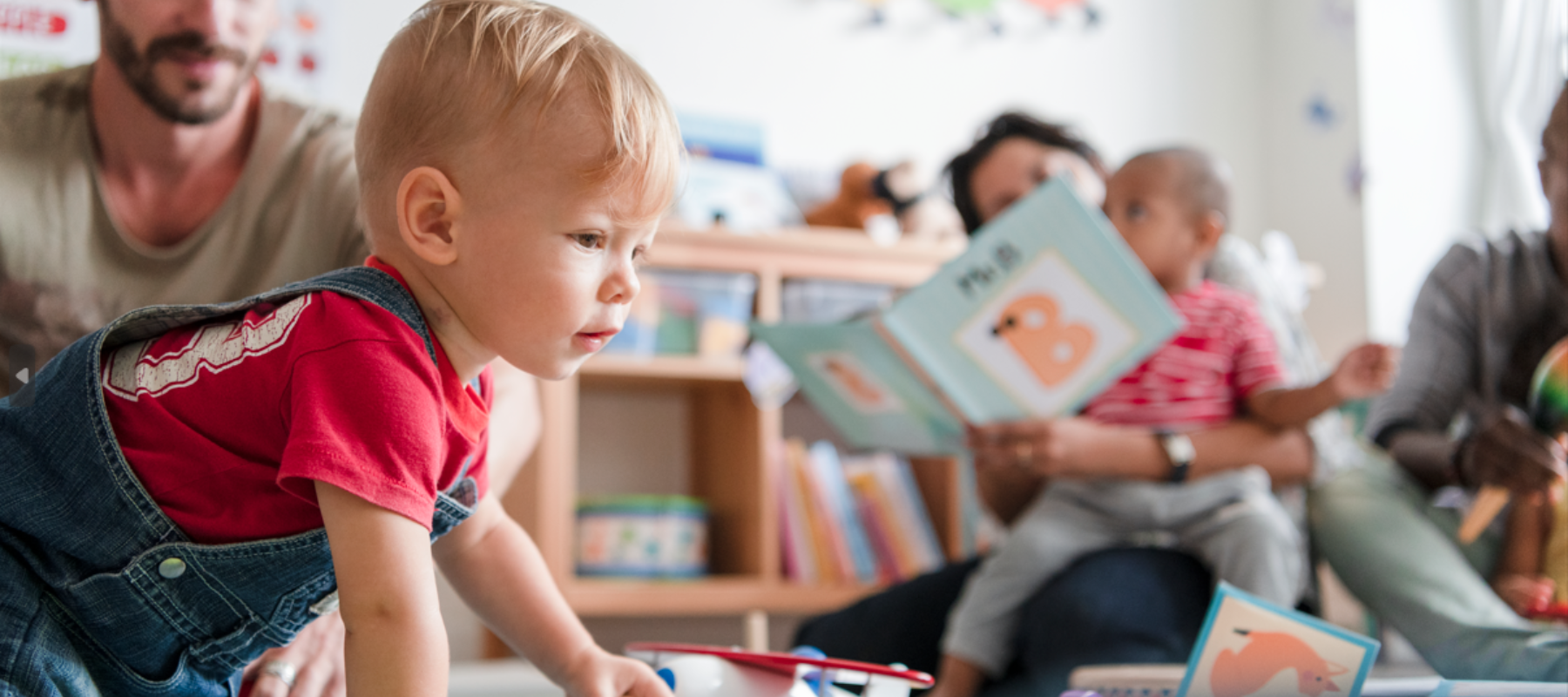 A toddler playing with an adult reading in the background