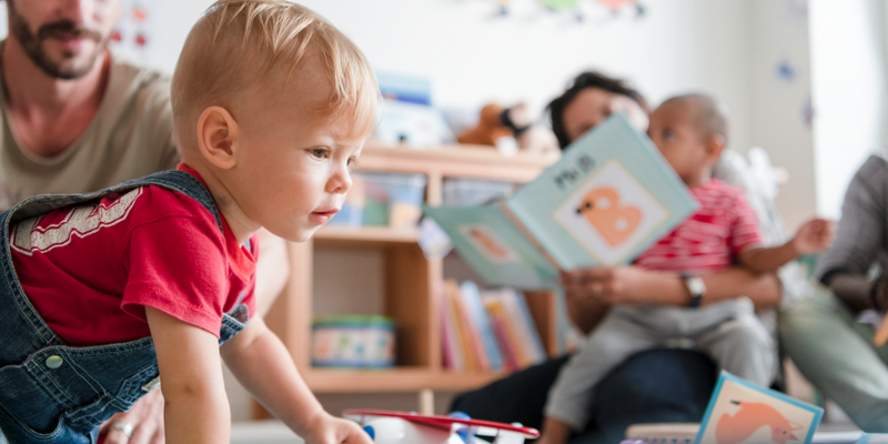 A toddler playing with a adult reading in the background