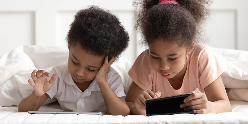 two children lying on a bed looking at tablets