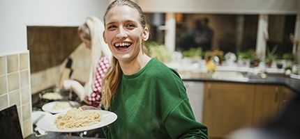 A young adult holding a plate of spaghetti and smiling whilst in a kitchen